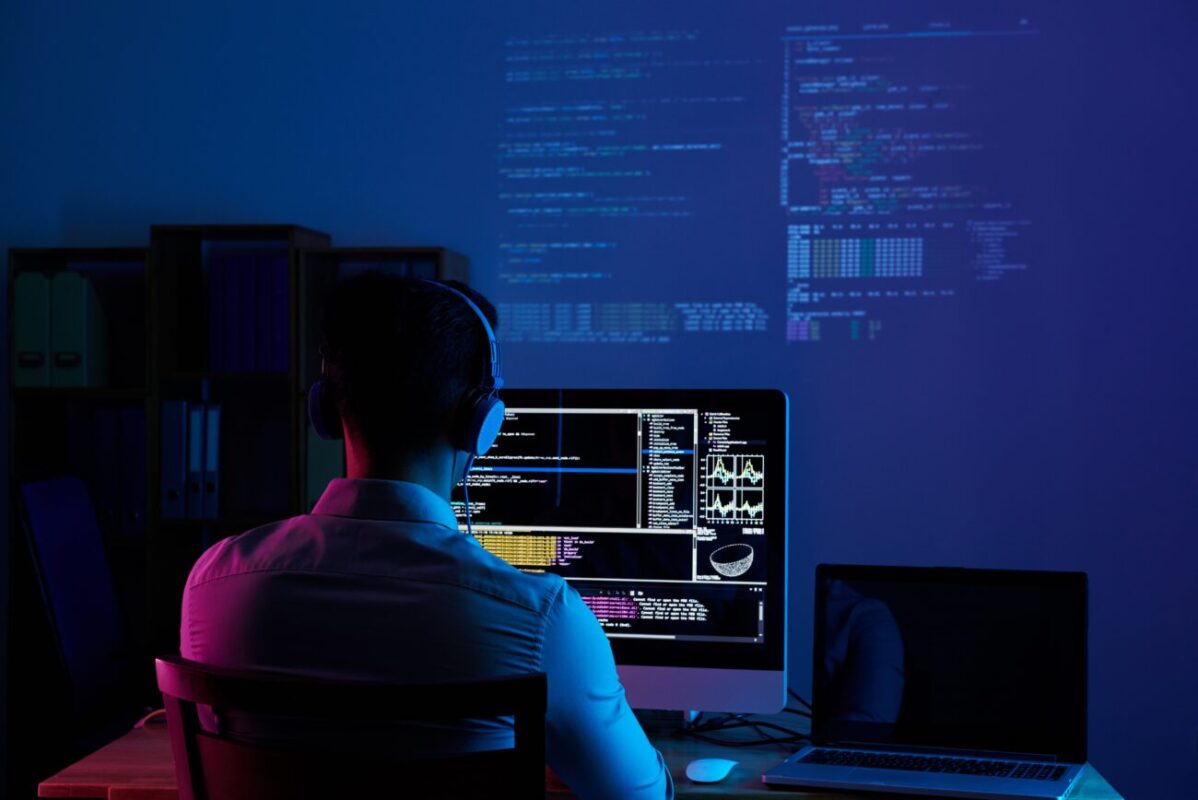 Person wearing headphones, seated at a desk coding on a large monitor with lines of code visible in a dark blue-lit room.
